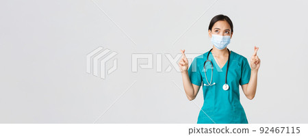Covid-19, coronavirus disease, healthcare workers concept. Hopeful serious-looking asian doctor, female physician in medical mask and scrubs cross fingers, looking upper left corner 92467115