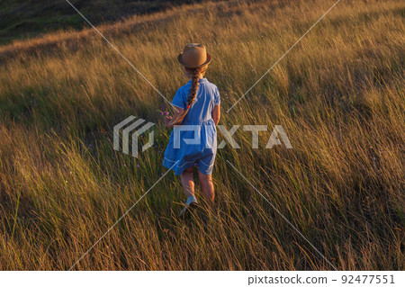 Adorable little girl in a straw hat, blue plaid summer dress in grass field countryside. Child with long blonde braid. Adorable little girl in a straw hat, blue plaid summer dress in grass field countryside. Child with long blonde braid. 92477551
