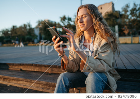 Young casual woman in coat sitting on bench on sea embankment using smartphone 92479096