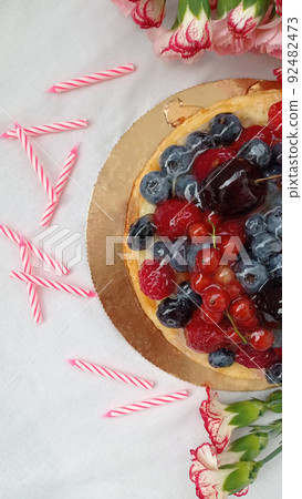 Berry cake. Cheesecake with cherries, raspberries, currants and blueberries. Birthday cake. Half of a cheesecake, pink candles and red flowers on a white background. Photo for stories. 92482473