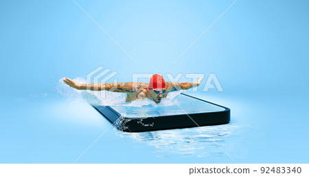 Creative collage with strong swimmer in goggles swimming by butterfly style in water in 3d phone screen over blue background. Sport, competition, online watching sports event Creative collage with strong swimmer in goggles swimming by butterfly style in water in 3d phone screen over blue background. Sport, competition, online watching sports event 92483340