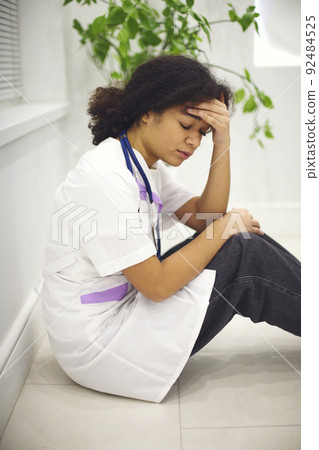 Sad tired young african american nurse sitting on floor in clinic, feeling overwhelmed by workload Sad tired young african american nurse sitting on floor in clinic, feeling overwhelmed by workload 92484525