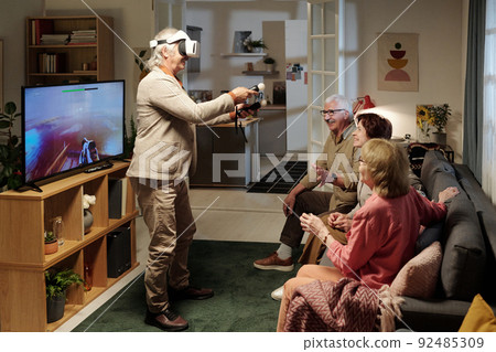 Group of cheerful senior friends looking at mature man with gamepads Group of cheerful senior friends looking at mature man with gamepads 92485309