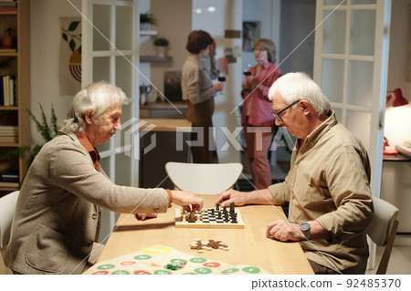Two friendly senior men sitting by table in living room and playing chess Two friendly senior men sitting by table in living room and playing chess 92485370