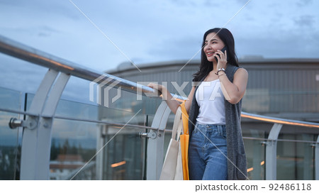 Stylish young woman talking on mobile phone while standing on rooftop terrace, overlooking the city view 92486118