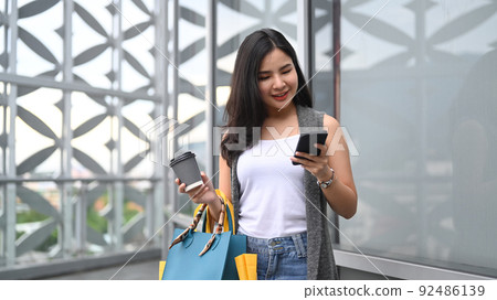 Smiling young woman with shopping bags, using smartphone and walking outside of shopping mall 92486139