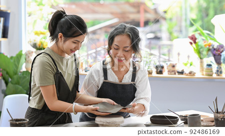 Senior female potter teaching young woman creating and sculpting clay pot in craft studio workshop 92486169