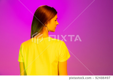 Portrait of young girl posing in casual white T-shirt isolated over pink background in neon light. Back view Portrait of young girl posing in casual white T-shirt isolated over pink background in neon light. Back view 92486497