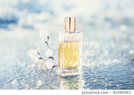 Close up of rectangular transparent glass bottle of golden perfume stands near snowy twig on a frosted pond Close up of rectangular transparent glass bottle of golden perfume stands near snowy twig on a frosted pond 92490963