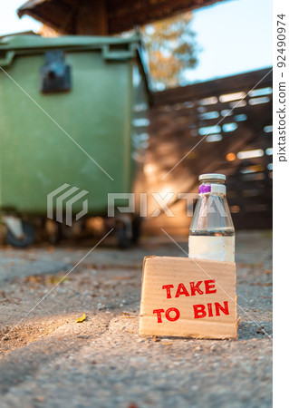 The concept of environmental pollution and waste sorting. A glass bottle stands on the ground in front of a dumpster. Near cardboard with the inscription TAKE to BIN. Vertical orientation The concept of environmental pollution and waste sorting. A glass bottle stands on the ground in front of a dumpster. Near cardboard with the inscription TAKE to BIN. Vertical orientation 92490974
