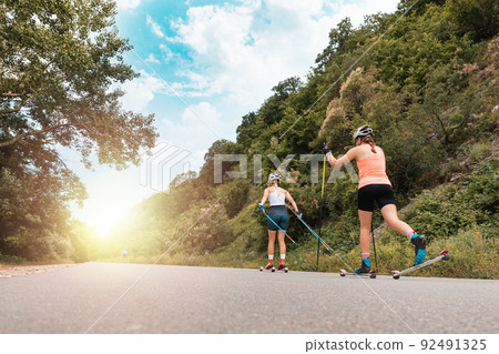 Two athletic women together training on the roller ski. Back low angle view. Copy space. Sunlight at finish. Concept of competition, biathlon, and summer workout 92491325