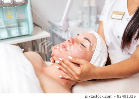 Cosmetologist makes a woman a facial massage procedure. Portrait of the client and the doctor's hands in close-up. An electronic device in the background. Concept of cosmetology and treatment Cosmetologist makes a woman a facial massage procedure. Portrait of the client and the doctor's hands in close-up. An electronic device in the background. Concept of cosmetology and treatment 92491329
