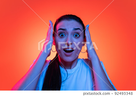 Young emotional stressed girl, student in white t-shirt looking at camera wide open eyes isolated on orange color background in neon light. 92493348
