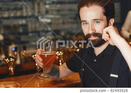Attractive young man standing behind the bar. Bearded man drinking beer. Attractive young man standing behind the bar. Bearded man drinking beer. 92493826