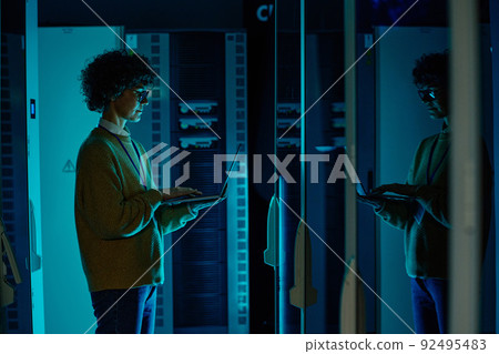 IT engineer standing before working server rack doing routine maintenance check and diagnostics using laptop 92495483