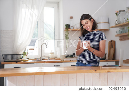 A woman in the kitchen with a smile. A woman drinks coffee and holds a cup in her hand. She is happy because She drinks her favourite coffee in the morning. High quality photo with copy space. A woman in the kitchen with a smile. A woman drinks coffee and holds a cup in her hand. She is happy because She drinks her favourite coffee in the morning. High quality photo with copy space. 92495930