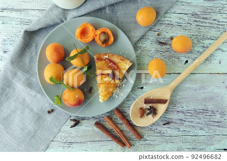 A piece of apricot pie on a plate, sprinkled with powdered sugar, spices and mint leaves, fresh apricots on the kitchen table, homemade food, sweet dessert A piece of apricot pie on a plate, sprinkled with powdered sugar, spices and mint leaves, fresh apricots on the kitchen table, homemade food, sweet dessert 92496682