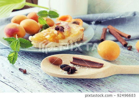 A piece of apricot pie on a plate, sprinkled with powdered sugar, spices and mint leaves, fresh apricots on the kitchen table, homemade food, sweet dessert A piece of apricot pie on a plate, sprinkled with powdered sugar, spices and mint leaves, fresh apricots on the kitchen table, homemade food, sweet dessert 92496683
