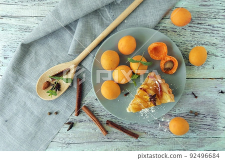 A piece of apricot pie on a plate, sprinkled with powdered sugar, spices and mint leaves, fresh apricots on the kitchen table, homemade food, sweet dessert A piece of apricot pie on a plate, sprinkled with powdered sugar, spices and mint leaves, fresh apricots on the kitchen table, homemade food, sweet dessert 92496684