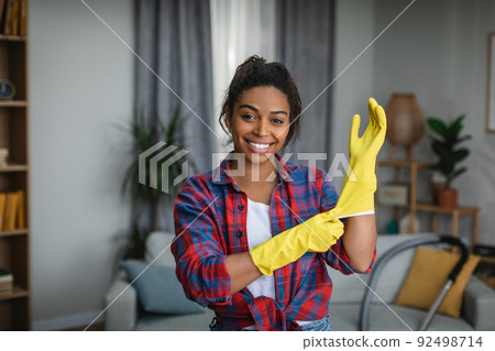 Smiling young african american woman puts on rubber gloves and ready to homework in living room interior Smiling young african american woman puts on rubber gloves and ready to homework in living room interior 92498714