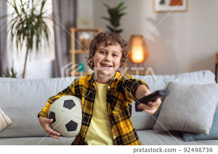 Little boy football fan watching tv at home, sitting on sofa with remote controller and soccer ball, smiling to camera 92498736