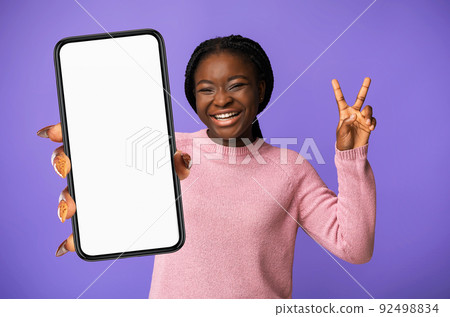 Joyful Black Young Woman Gesturing V-Sing And Showing Smartphone, Mockup 92498834