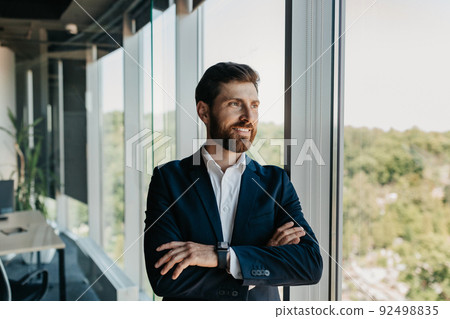 Portrait of handsome caucasian businessman standing with folded arms near window, having break in office, free space 92498835