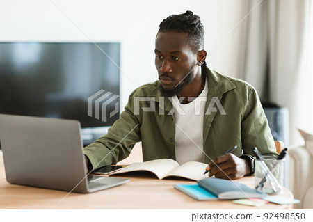 Focused african american man using laptop, working remotely and taking notes, having remote meeting from home 92498850