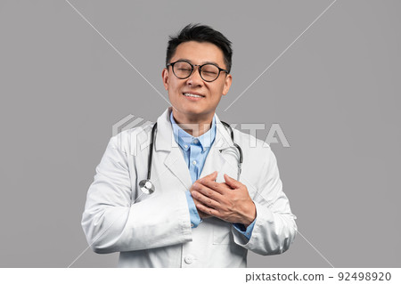 Happy millennial korean guy doctor in white coat, glasses with stethoscope presses hands to heart Happy millennial korean guy doctor in white coat, glasses with stethoscope presses hands to heart 92498920