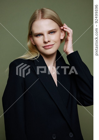 A young charming blonde model tucks her hair behind her ears touches and strokes her curls posing on a green background in the studio A young charming blonde model tucks her hair behind her ears touches and strokes her curls posing on a green background in the studio 92499896