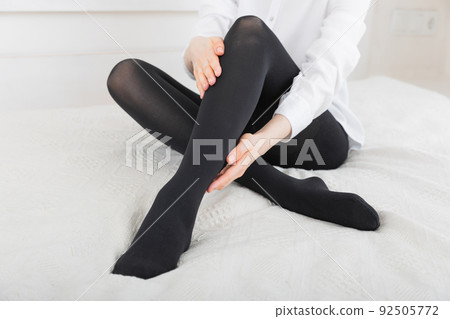 Black compression stockings on a woman in a white room. Black tights. Girl putting on stockings at home. Beautiful female legs. 92505772