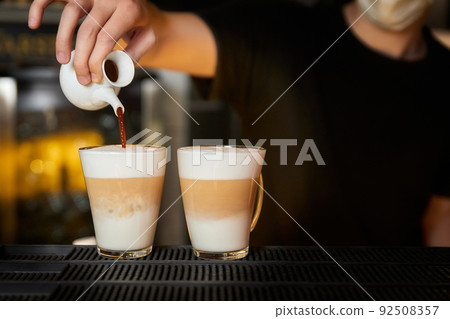 Bartender prepares coffee latte by pouring Black coffee into a glass mug with frothed milk Bartender prepares coffee latte by pouring Black coffee into a glass mug with frothed milk 92508357