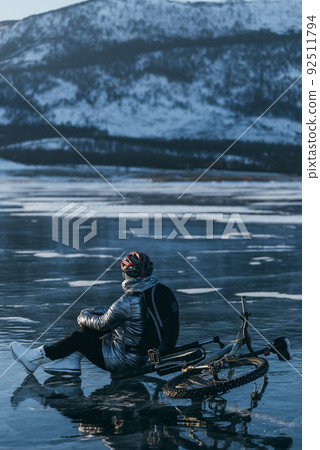 Woman sitting near her bike on ice. The girl cyclist stopped to rest. She sits on the wheel and enjoys the beautiful view of the sunset. Woman sitting near her bike on ice. The girl cyclist stopped to rest. She sits on the wheel and enjoys the beautiful view of the sunset. 92511794
