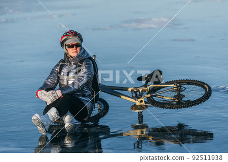 Woman sitting near her bike on ice. The girl cyclist stopped to rest. She sits on the wheel and enjoys the beautiful view of the sunset. 92511938