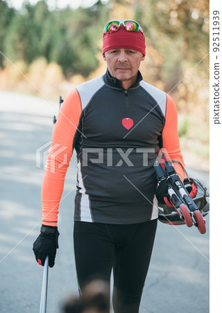 Training an athlete on the roller skaters. Biathlon ride on the roller skis with ski poles, in the helmet. Autumn workout. Roller sport. The athlete goes and holds sports equipment in his hand. Training an athlete on the roller skaters. Biathlon ride on the roller skis with ski poles, in the helmet. Autumn workout. Roller sport. The athlete goes and holds sports equipment in his hand. 92511939