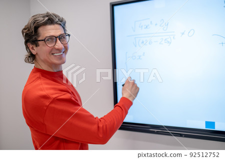 A teacher in red shirt writing on the board at the lesson and looking contented 92512752