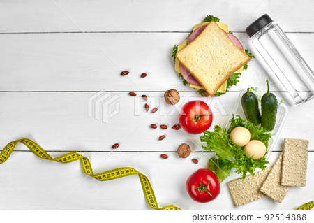 Lunch boxes with sandwich and fresh vegetables, bottle of water, nuts and eggs on white wooden background. Top view with copy space Lunch boxes with sandwich and fresh vegetables, bottle of water, nuts and eggs on white wooden background. Top view with copy space 92514888
