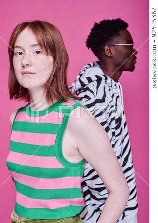 Portrait of red haired teenage girl looking at camera with African teenage boy standing behind her over pink background 92515362