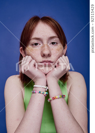 Portrait of red haired teenage girl with handmade bracelets on her arms looking at camera on blue background Portrait of red haired teenage girl with handmade bracelets on her arms looking at camera on blue background 92515429