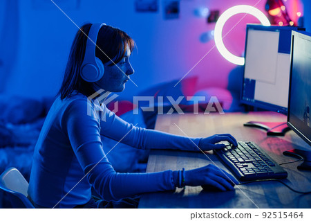 Teenage girl in wireless headphones playing video game on computer while sitting at table in dark neon room Teenage girl in wireless headphones playing video game on computer while sitting at table in dark neon room 92515464