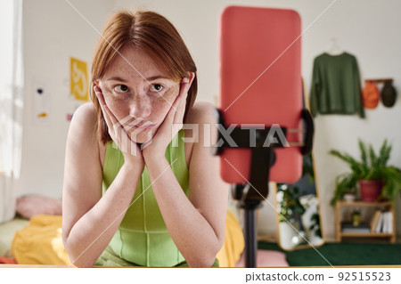 Teenage girl sitting at table in front of the smartphone on tripod and waiting for the connection of live broadcast Teenage girl sitting at table in front of the smartphone on tripod and waiting for the connection of live broadcast 92515523