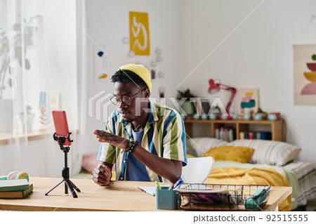 African young blogger sitting at table in front of smartphone on tripod and talking online with his followers 92515555
