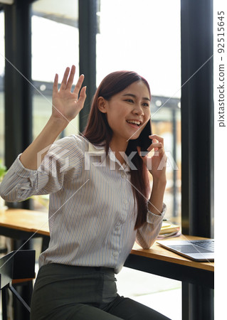 Cheerful female employee waving hands to greet her colleagues while talking on mobile phone Cheerful female employee waving hands to greet her colleagues while talking on mobile phone 92515645