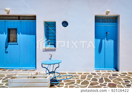 Traditional blue doors and windows and whitewashed walls of Greek houses. Midday summer sunshine, blue table, cobblestone pavement, Milos, Greece 92516422