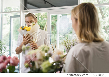 Young bearded man buys a beautiful bouquet of flowers for a girl's holiday in a cozy flower shop. 92518350