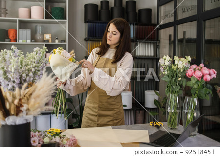 Portrait of young brunette female florist holding bouquet of flowers and looking at them. 92518451