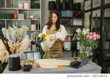 Portrait of young brunette female florist holding bouquet of flowers and looking at them. 92518453