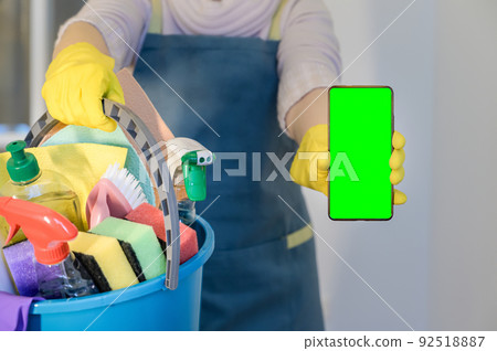 homeowner in rubber gloves holding detergent bottles. Hold mobile phone with blank empty green screen 92518887