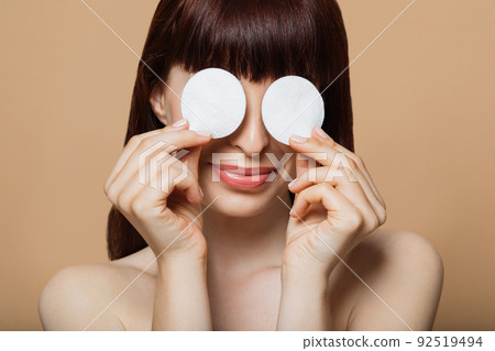 Smiling young woman covering eyes with two cotton pads that holding in hands over beige background 92519494