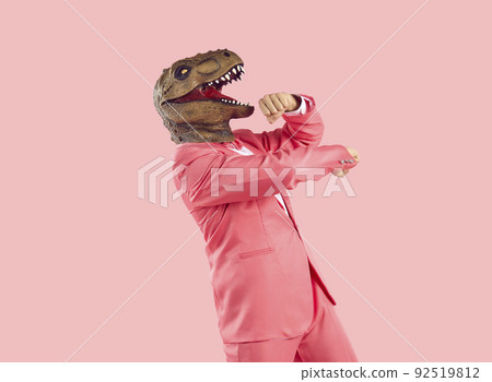 Cheerful funny and humorous man in dinosaur rubber mask dances isolated on pastel pink background. 92519812